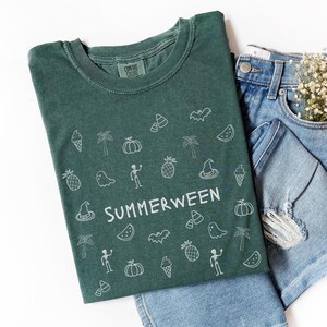 May include: A green t-shirt with the word "SUMMERWEEN" in white, surrounded by white line art of Halloween and summer-themed icons. The shirt is paired with denim shorts and a sprig of white flowers.