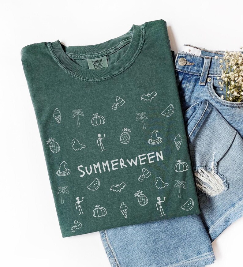 May include: A green t-shirt with the word "SUMMERWEEN" in white, surrounded by white line art of Halloween and summer-themed icons. The shirt is paired with denim shorts and a sprig of white flowers.