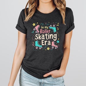 May include: A dark heather grey t-shirt with a graphic design featuring roller skates, stars, and music notes. The text on the shirt reads "In my Roller Skating Era".