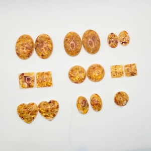 Peut inclure: Une collection de cabochons en résine de couleur ambre de différentes formes. L'assortiment comprend des pièces ovales, rondes, carrées et en forme de cœur, avec un aspect moucheté et texturé. Ces cabochons conviennent à la fabrication de bijoux et à l'artisanat.