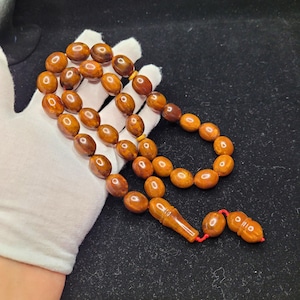 May include: A string of amber-coloured prayer beads with oval-shaped beads. The beads are strung together with a red cord and feature a decorative tassel. The beads are held by a gloved hand against a dark background.