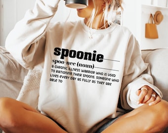 May include: A white sweatshirt with the definition of the word "spoonie" printed on it. The definition reads: "A chronic illness warrior who is used to rationing their spoons. Someone who lives every day as fully as they are able to."
