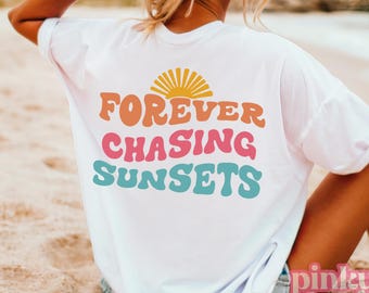 May include: White t-shirt with a retro-style graphic that says "FOREVER CHASING SUNSETS" in rainbow colors. The shirt is being worn by a person sitting on a sandy beach.