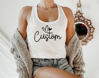 May include: White tank top with a black graphic of two hearts and the word "Custom" on the front. The tank top is being worn by a person with a light gray cardigan.