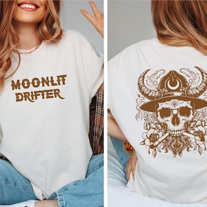 May include: A white t-shirt with brown text that reads "MOONLIT DRIFTER" on the front and a brown illustration of a skull wearing a hat with horns and a crescent moon on the back.