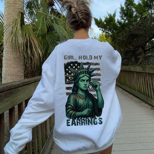 May include: White sweatshirt with a graphic of the Statue of Liberty holding an earring, with the text "GIRL, HOLD MY EARRINGS" in a distressed font. The design incorporates an American flag motif. The sweatshirt is worn outdoors.