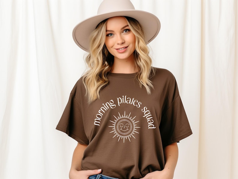 May include: A brown t-shirt with a white graphic of a sun with a smiling face and the text "morning pilates squad" around it.