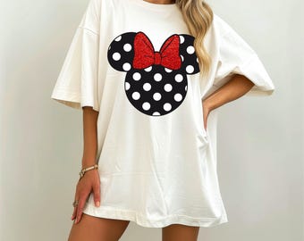 May include: Cream-coloured oversized t-shirt featuring a black and white polka-dotted mouse head design with a red glitter bow. The shirt has short sleeves and a relaxed fit, suitable for casual wear.