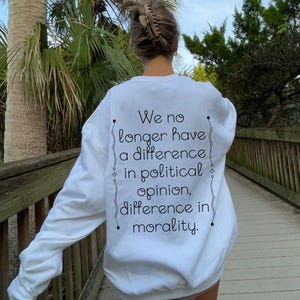May include: White sweatshirt with the text "We no longer have a difference in political opinion, difference in morality" in black script. The sweatshirt is worn by a person walking on a wooden boardwalk, with palm trees and greenery in the background.
