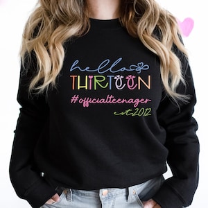 May include: Black sweatshirt with the words "hello THIRTEEN #officialteenager est. 2012" in colorful script. The text is in a rainbow of colors, including blue, yellow, pink, and green. Pink heart decorations are in the background.
