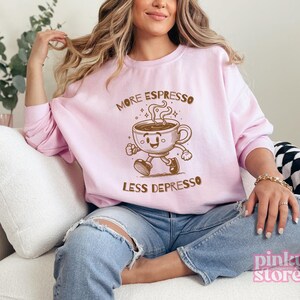 May include: A light pink sweatshirt with the words "MORE ESPRESSO LESS DEPRESSO" printed in brown. The graphic features a cartoon coffee cup with a smiling face and legs. The sweatshirt is a casual, comfortable style.