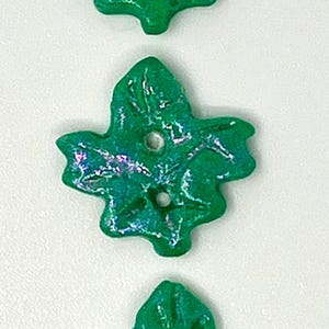 Peut inclure: Trois boutons verts en forme de feuille avec des reflets irisés. Chaque bouton comporte deux trous pour la couture. Les boutons sont disposés verticalement sur un fond blanc, mettant en valeur leur design et leur couleur uniques.