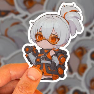 May include: A sticker featuring a chibi-style anime character with white hair and orange goggles. The character wears a black and orange outfit with a backpack. The sticker is held by a hand.