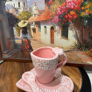 May include: A pink ceramic teacup and saucer set with a matching spoon. The set features a white floral design. The teacup is heart-shaped. The set is displayed on a wooden surface, with a framed painting in the background.