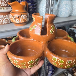 May include: Hand-held terracotta serving dish with four bowls and a central holder. The bowls are decorated with white and green floral patterns. Other terracotta pottery items are visible in the background.
