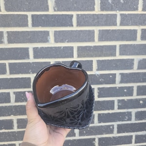 May include: A black ceramic heart-shaped mug with a brown interior and intricate floral designs. The mug is held in a hand.