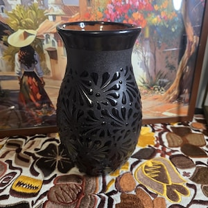 May include: A black vase with a textured surface and a floral pattern. The vase has a wide opening and a dark brown interior. The vase is sitting on a patterned tablecloth, with a painting in the background.