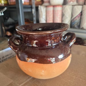 May include: A handcrafted, small, round, ceramic pot with two handles. The top half is a dark brown color, and the bottom half is a light orange color. The pot has a glossy finish and is likely used for decorative purposes.
