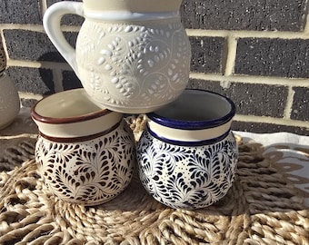 May include: Three ceramic mugs with different designs. The mugs are white with brown and blue floral patterns. The mugs are sitting on a woven rug.