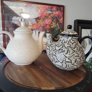 May include: Two ceramic teapots on a wooden tray. One is cream-colored with a leaf pattern, the other is black and white with a floral design. The tray has a dark rim with handwritten text.