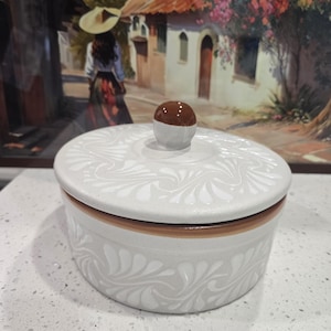 May include: A round, lidded ceramic container with a white and brown color scheme. The container features a decorative floral pattern and a small, round knob on the lid. The container is sitting on a white surface.