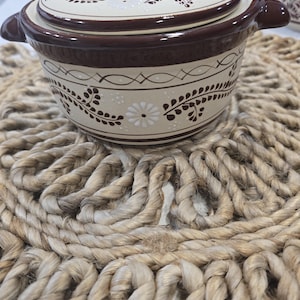 May include: A white ceramic casserole dish with a brown lid and floral design. The dish has a handle on the lid and two handles on the sides. The dish is sitting on a woven rug.
