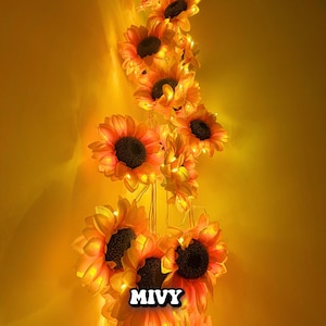 May include: A string of artificial sunflowers with warm white LED lights. The sunflowers have yellow petals with orange tips and dark brown centers. The string of flowers is against a yellow background. The text "MIVY" is at the bottom.