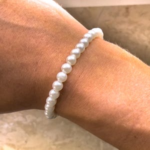 Könnte beinhalten: Ein weißes Perlenarmband mit schlichtem Design. Das Armband besteht aus kleinen, runden Perlen, die auf einer durchsichtigen Gummikordel aufgefädelt sind.