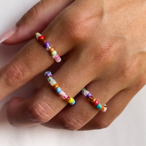 Könnte beinhalten: Drei bunte Perlenringe an einer Hand. Die Ringe bestehen aus kleinen, runden Perlen in verschiedenen Farben, darunter Rosa, Orange, Gelb, Grün, Blau, Lila und Weiß.