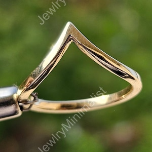 Puede incluir: Un anillo dorado con un diseño en forma de V. El anillo está sujeto por una herramienta plateada sobre un fondo verde borroso. El diseño del anillo es minimalista y moderno, con un acabado pulido.