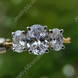 May include: A three-stone ring with three oval gemstones. The central stone is larger than the two side stones. The ring has a gold band. The ring is held by a tool against a blurred green background.