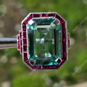Peut inclure: Gros plan sur une bague rectangulaire avec une grosse pierre centrale taillée en émeraude vert clair. La bague est entourée d'une bordure de pierres magenta plus petites et rectangulaires. La bague est tenue par un outil argenté. Le texte "JewlryMinimalist" est visible.