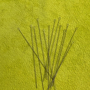 Könnte beinhalten: Ein Satz von 18 dünnen, silberfarbenen Reinigungsnadeln aus Metall mit einer Schlaufe oben. Die Nadeln sind fächerförmig auf einer grünen, flauschigen Oberfläche angeordnet.