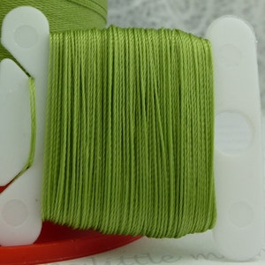 May include: A spool of green thread wound around a white plastic bobbin with two holes. The thread is smooth and appears to be made of cotton.