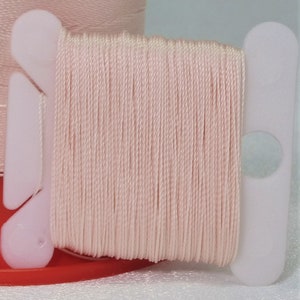 Puede incluir: Una bobina de hilo rosa pálido enrollada en una bobina de plástico blanca. El hilo es liso y parece ser de algodón.