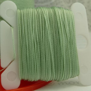 May include: A spool of light green thread, tightly wound and displayed on a white plastic holder. The thread is a soft, muted green color, and the spool is set against a blurred background.