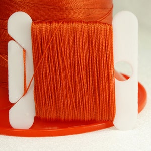 May include: A spool of orange thread wound around a white plastic bobbin with two white plastic thread guides. The thread is wound tightly and evenly around the bobbin.