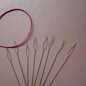 May include: A set of ten silver threader needles with a loop at the end. The needles are arranged in a fan shape on a light pink surface. A circle of pink thread is in the upper left corner of the image.