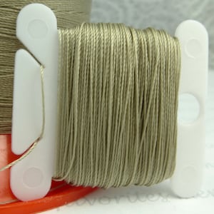 Può includere: Un'bobina di filo di colore marrone chiaro avvolta attorno a un porta-bobina di plastica bianca. Il filo è liscio e ha una lucentezza sottile.