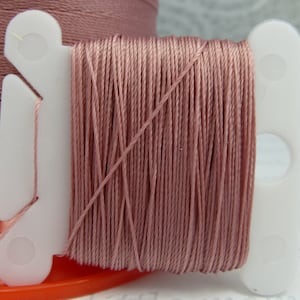 May include: A spool of dusty rose colored thread wound on a white plastic thread holder.