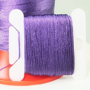 May include: A spool of purple thread with white plastic bobbins on either side. The thread is wound around a red plastic base.