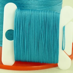 May include: A spool of teal blue thread wound around a white plastic bobbin. The thread is held in place by a white plastic spool holder.