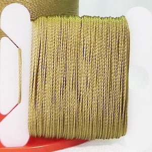 Op de afbeelding: Een spoel met goudkleurig garen gewikkeld om een witte plastic spoel. Het garen is dun en lijkt van synthetisch materiaal te zijn.