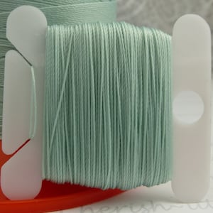 May include: A spool of light blue thread wound on a white plastic bobbin. The thread is smooth and silky.