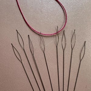 May include: A set of ten thin, silver-colored, needle-like threaders with a loop at the top. A single strand of pink thread is looped around the top of the threaders.