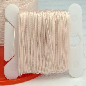 May include: A spool of pale pink thread wound on a white plastic bobbin with a white plastic holder.