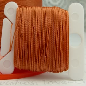 May include: A spool of burnt orange embroidery floss wound on a white plastic bobbin. The floss is ready to be used for crafting projects.