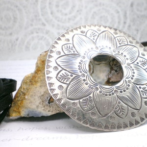 May include: A silver pendant with a detailed floral design. The pendant is round and has a hole in the center. It is sitting on a rock with a black leather cord.
