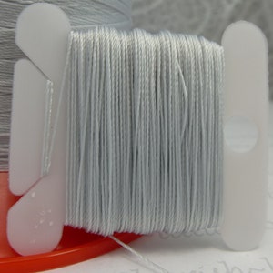 May include: A spool of light gray thread is the focal point of this image. The thread is tightly wound on a white plastic spool. The spool is set on a red base. Another spool of thread is visible in the background.