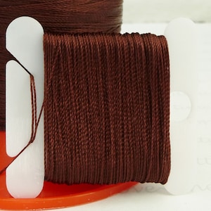 May include: A spool of brown embroidery floss wound around a plastic bobbin. The floss is held in place by two white plastic clips.